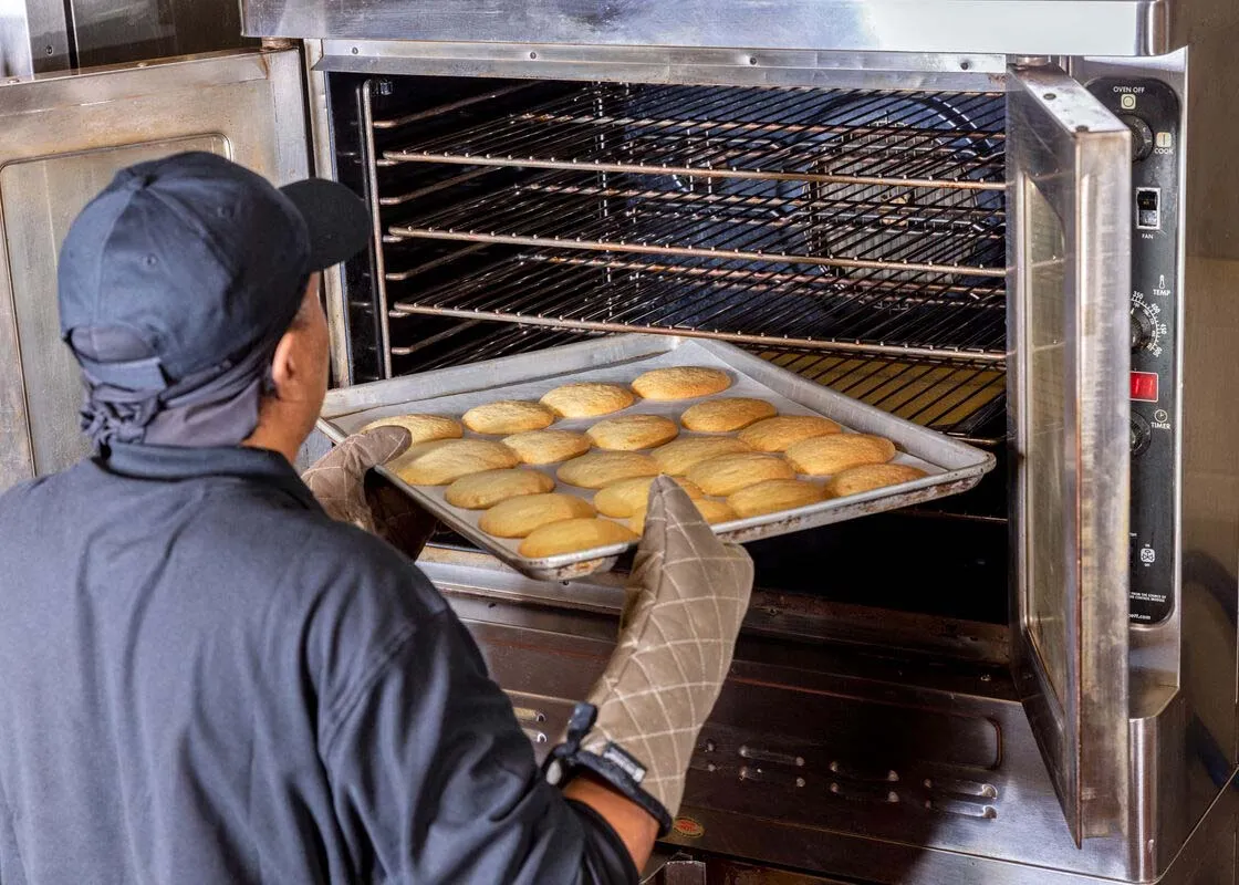A person wearing a black cap and oven mitts is taking a tray of freshly baked bread rolls out of a commercial oven in a kitchen setting.