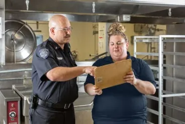 A police officer and a woman are reviewing a document together in an industrial kitchen setting.