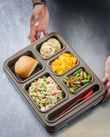A person holding a compartmentalized tray containing a bread roll, a cinnamon roll, mandarin orange slices, a mixed vegetable stir-fry, and a grain salad, with a red plastic fork placed in an empty section.