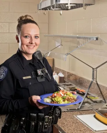 A smiling female police officer holding a plate of food in what appears to be a cafeteria or food service area.