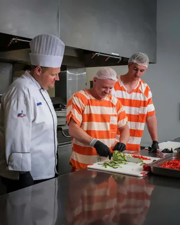 In a kitchen, a chef in a white uniform watches two men in orange-and-white striped prison uniforms as they prepare sliced vegetables on a cutting board.