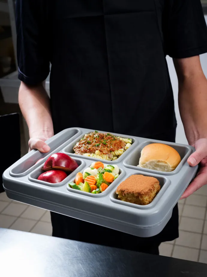 The image shows a person holding a compartmentalized tray with a meal consisting of a piece of lasagna, a bread roll, mixed vegetables, a breaded item, and sliced red apples.