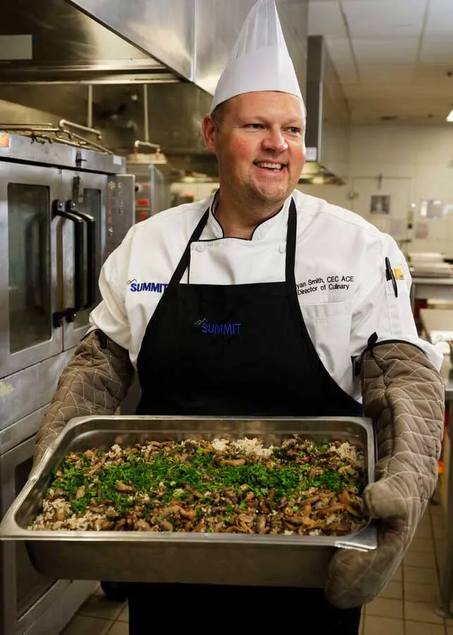 Smiling chef in a professional kitchen holding a large pan of plant based mushroom and lentil rice pilaf.